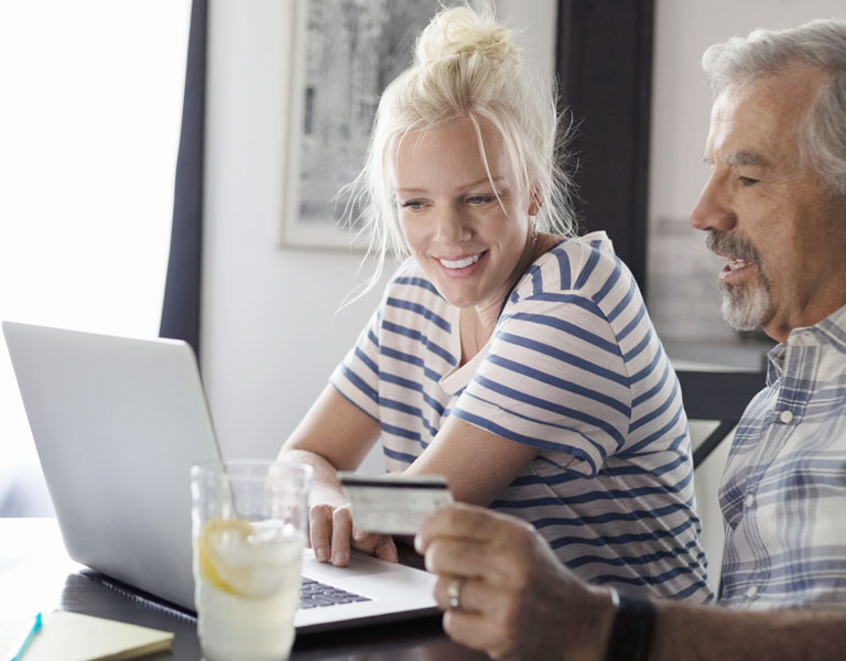 Um homem e uma mulher olhando sorridentes para a tela de um notebook. O homem segura um cartão de crédito.