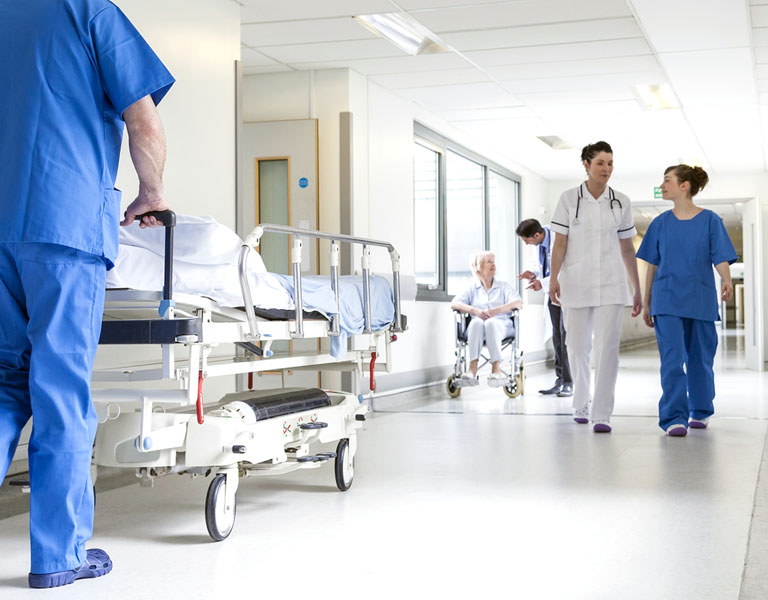 A imagem mostra um corredor de hospital. Um enfermeiro de roupas azuis empurra uma maca, uma médica de branco e uma enfermeira de azul conversam e uma senhora, sentada em uma cadeira de rodas, parece ser instruída por um médico. 