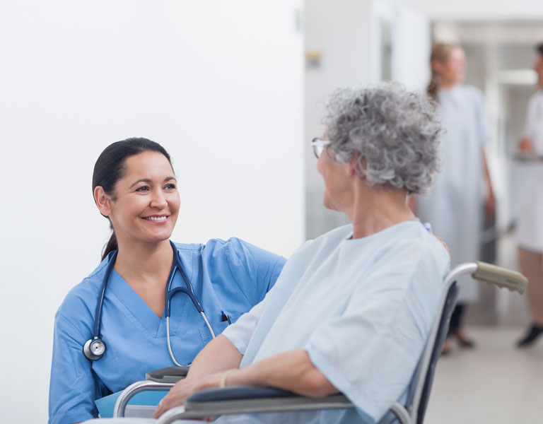 Enfermeira, num ambiente hospitalar, sorrindo enquanto parece conversar com uma paciente idosa que está na cadeira de rodas.