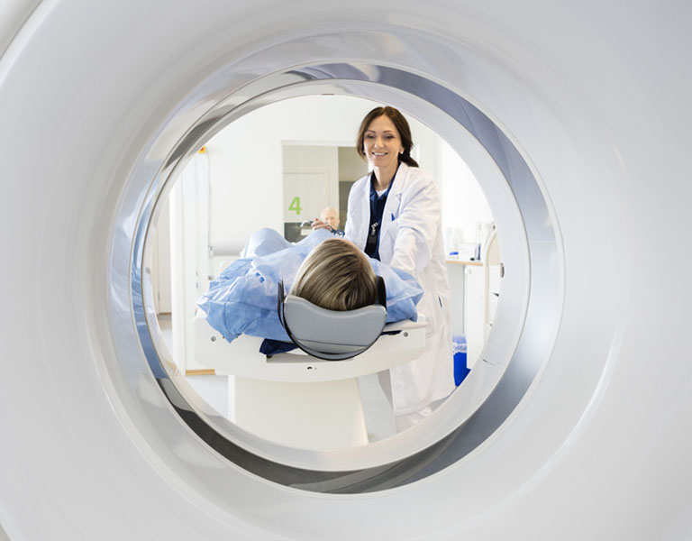 A imagem mostra uma médica de cabelos escuros sorrindo enquanto posiciona uma paciente para realizar uma ressonância magnética.